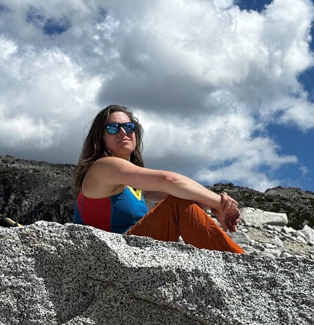 Kris Nóva sitting on a gray rock at the top of mountain with a blue sky and large cloud behind her.